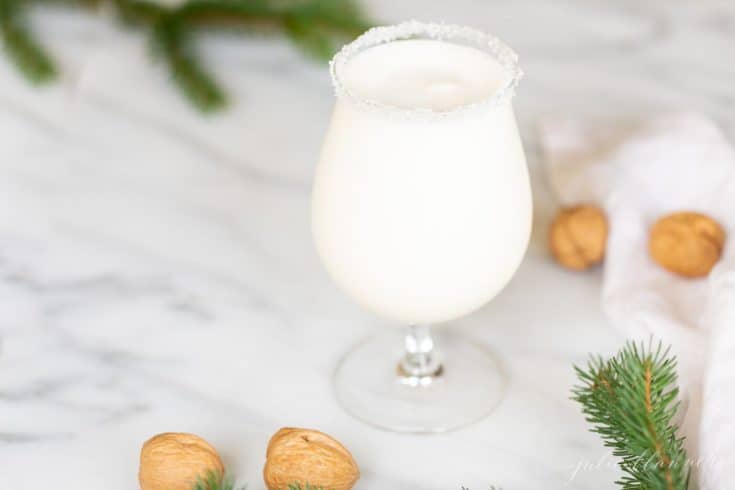 A clear stemmed glass filled with a white Christmas cocktail, topped with a sugared rim, hazelnuts and greenery to the side.