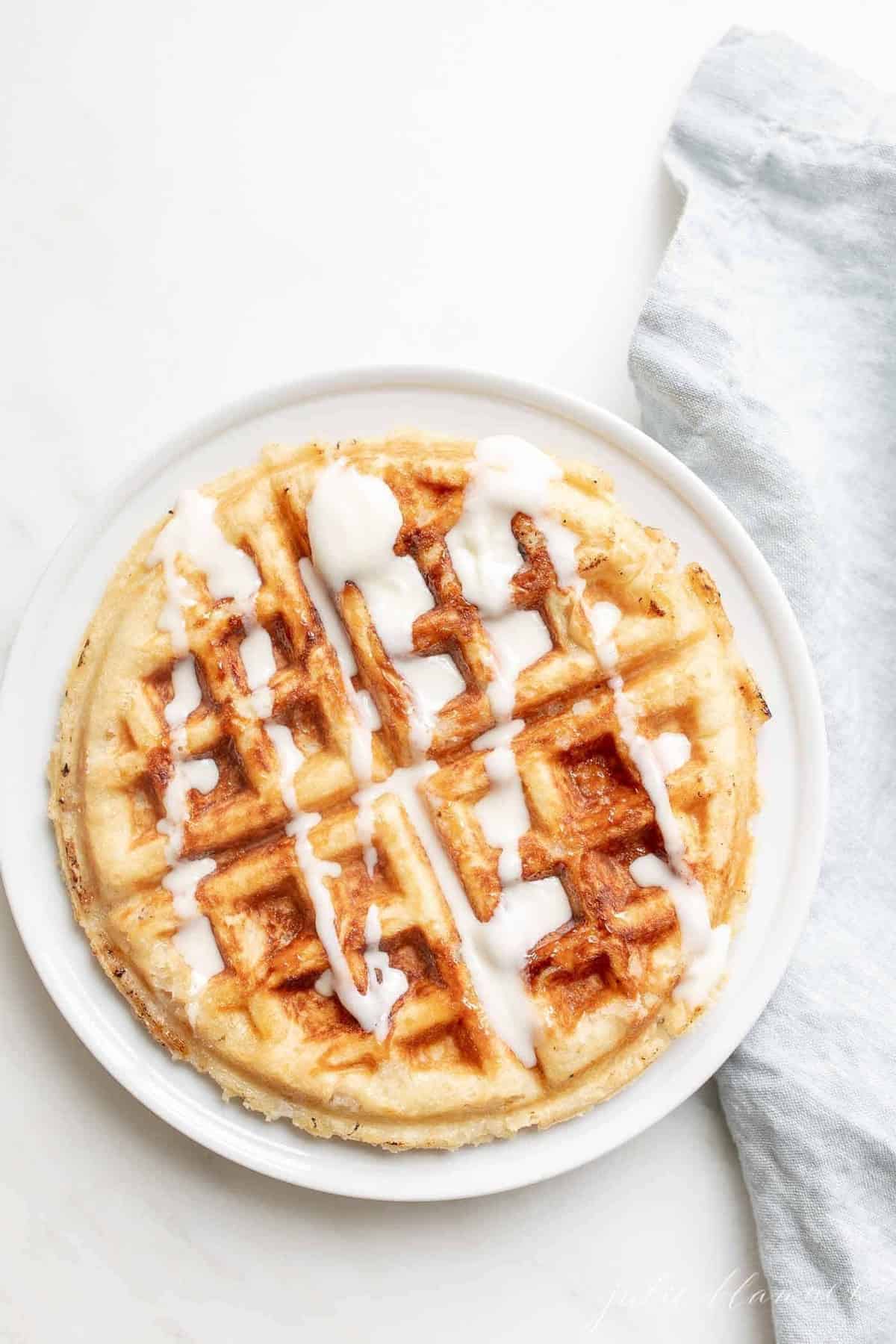 A Belgian waffle on a white plate, creme fraiche drizzled on top on a white plate. A blue linen napkin is to the side. 