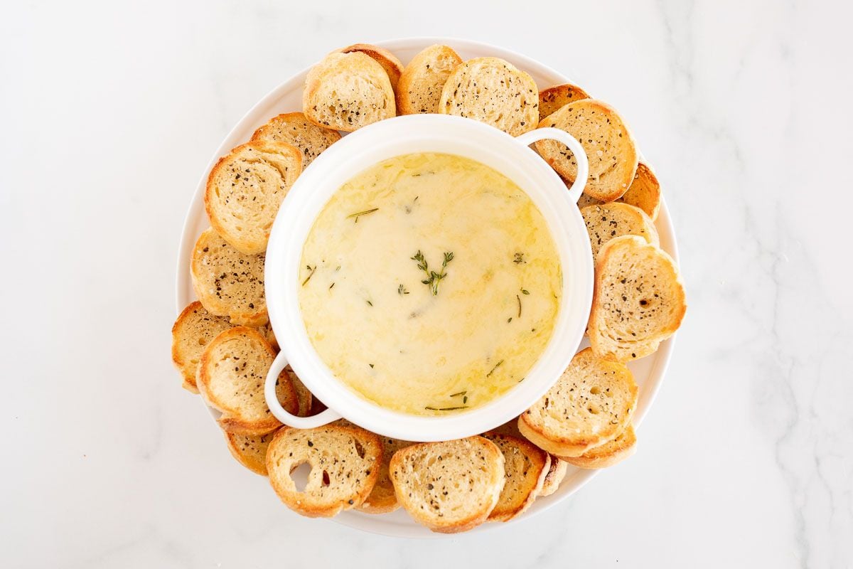 A white bowl of baked fontina cheese dip surrounded by crostini
