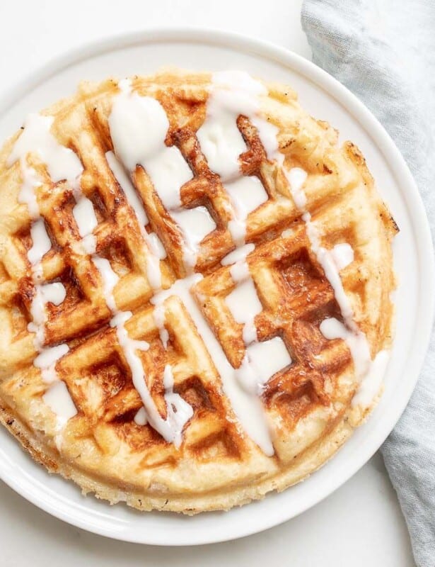A Belgian waffle topped with creme fraiche drizzle, on a white plate with a linen napkin to the side.