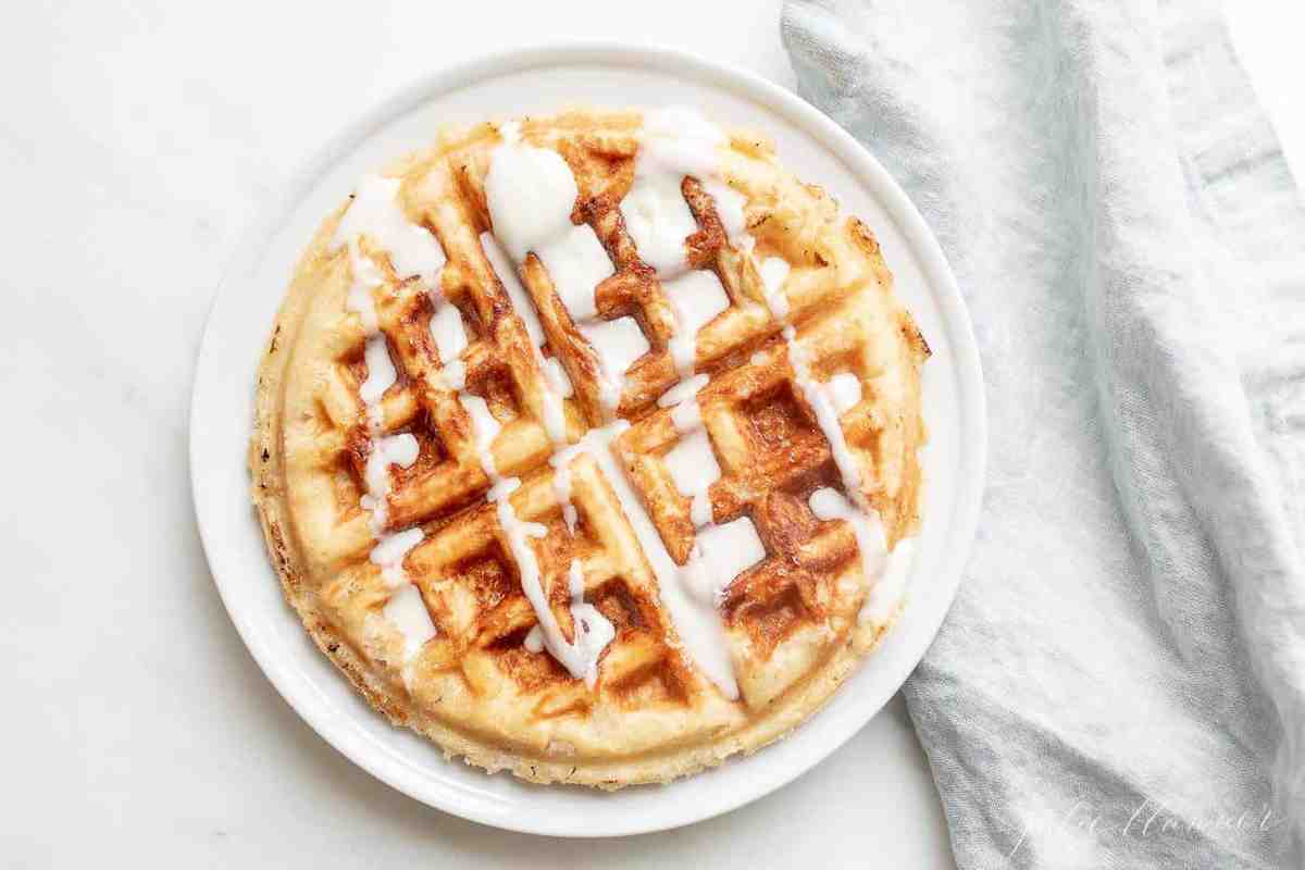 A Belgian waffle topped with creme fraiche drizzle, on a white plate with a linen napkin to the side. 