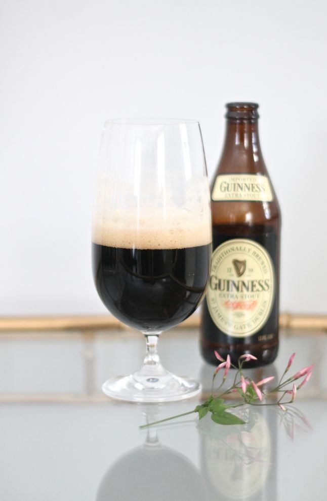 Bottle of Guinness wine next to a wine glass full, with a small sprig of purple jasmine on the table. 