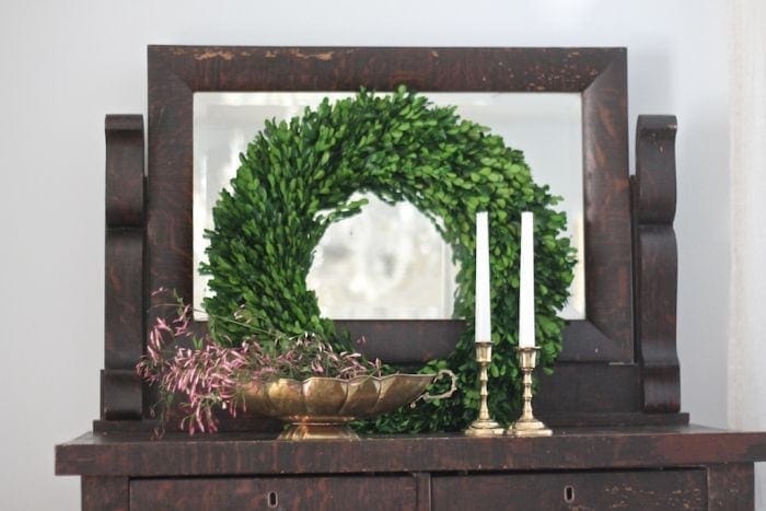 Vintage dresser top with boxwood wreath leaning in front of mirror, brass pedestal bowl filled with purple jasmine, and two brass candlesticks.