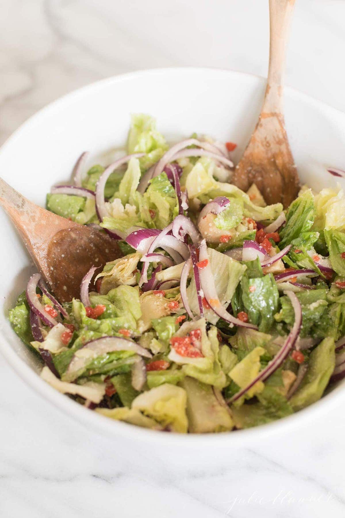 italian salad mixed up and topped with parmesan in a white bowl, with wooden serving spoons.