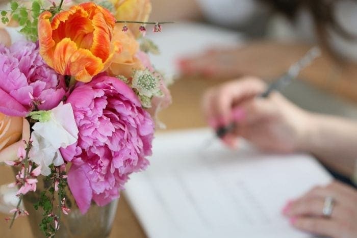 A bouquet of flowers in a vase on a table with someone practicing calligraphy in the background.