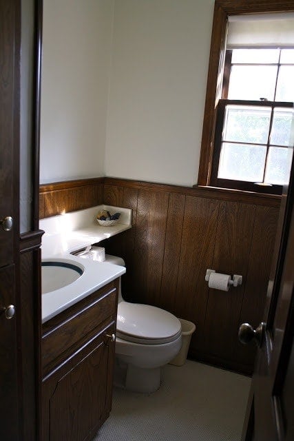 A dark and dated wood paneled half bathroom in a "before" photo of a renovation.