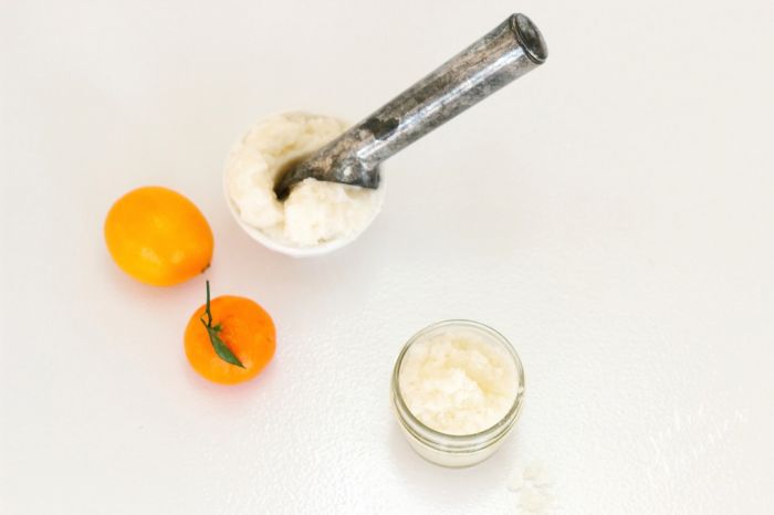 A white surface with small oranges and a scoop for a glass jar of sugar scrub.