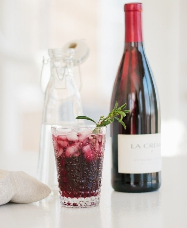 A clear glass featuring a red wine spritzer, with a bottle of wine in the background