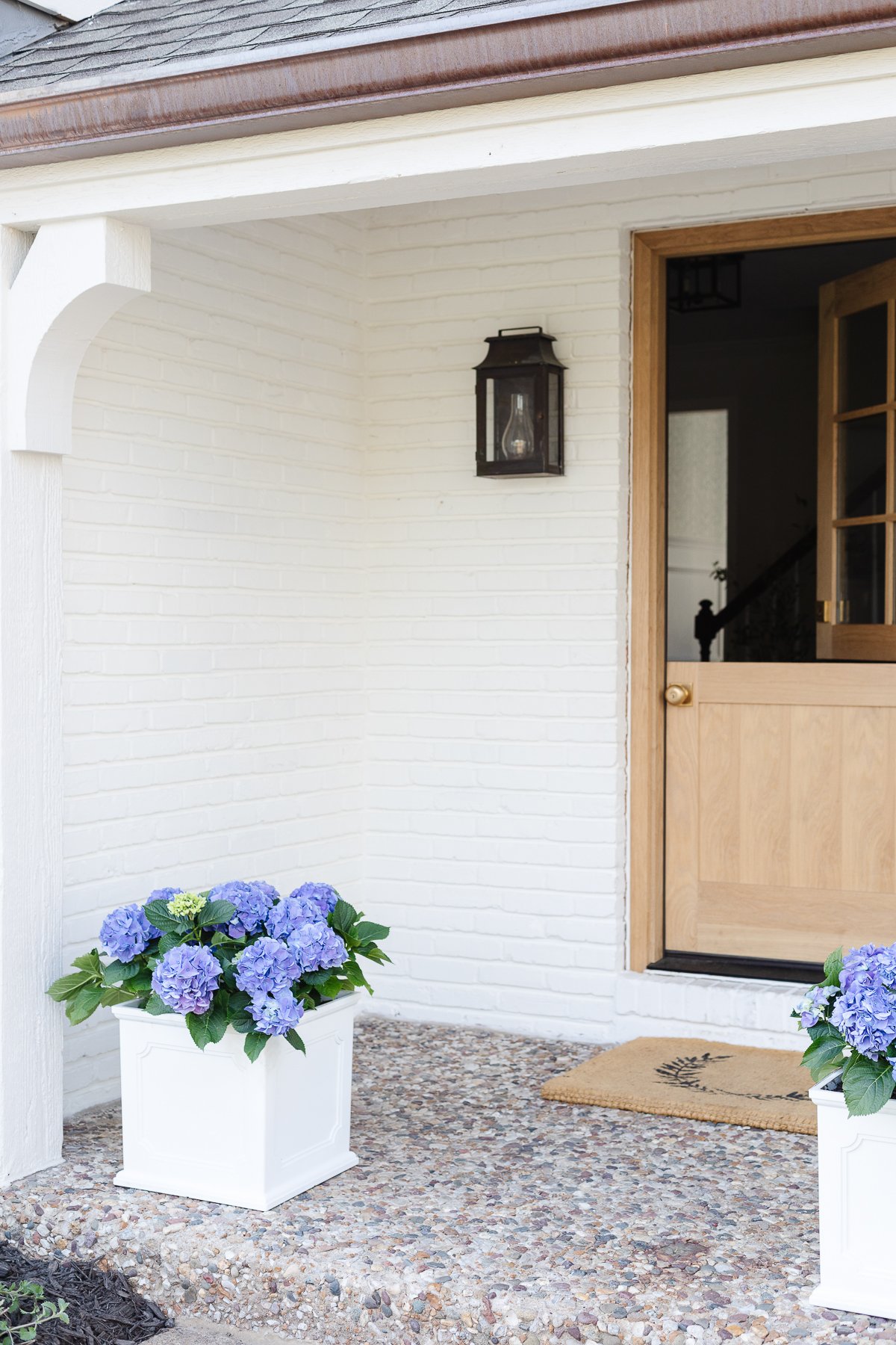 A white brick house with a wooden Dutch door and white planters on the spring porch, filled with blue hydrangeas.