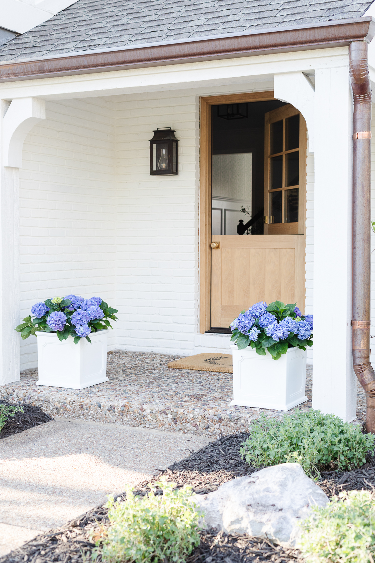 A white brick house with a wooden Dutch door and white planters on the spring porch, filled with blue hydrangeas.