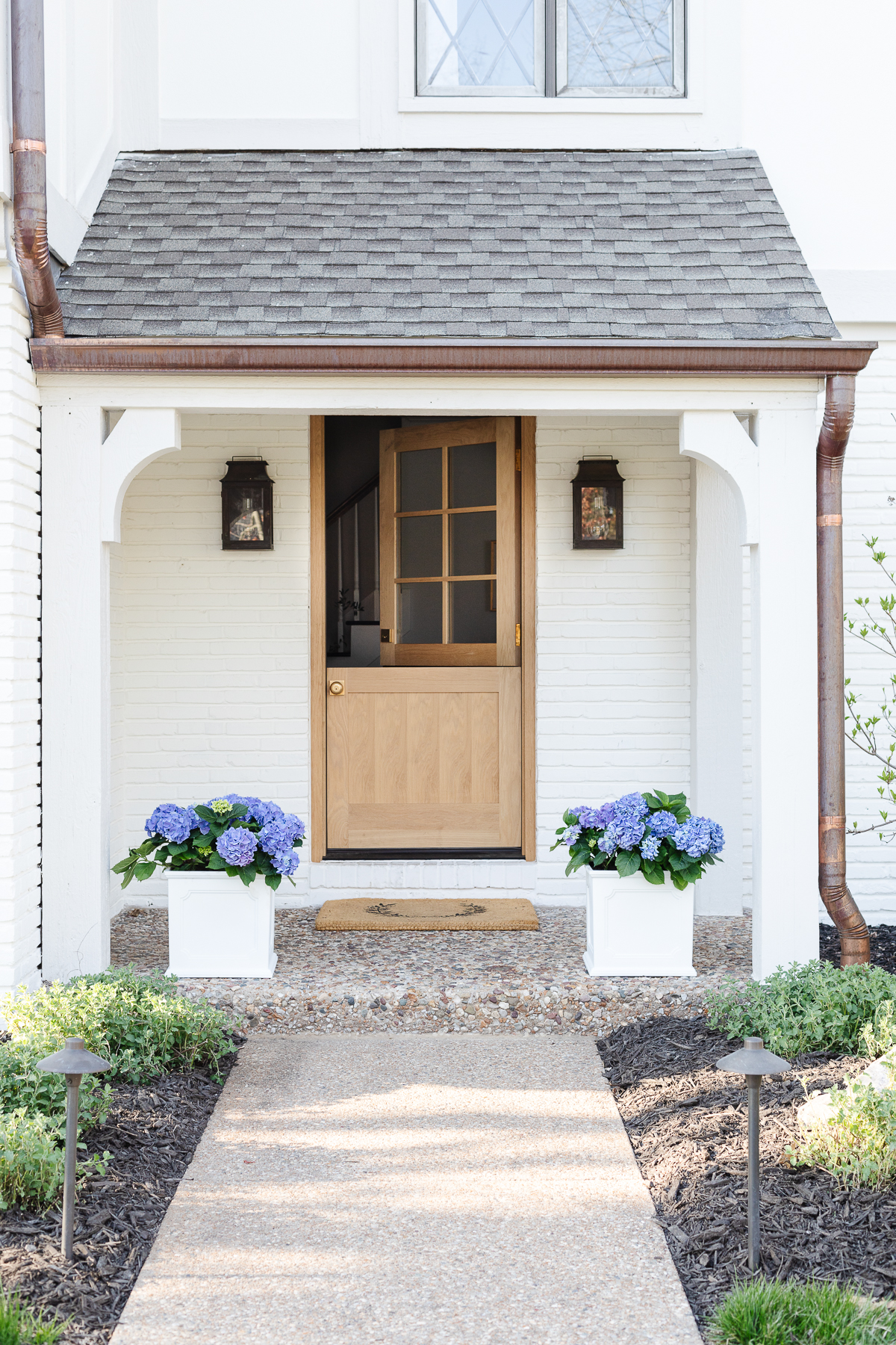 A white brick house with a wooden Dutch door and white planters on the spring porch, filled with blue hydrangeas.