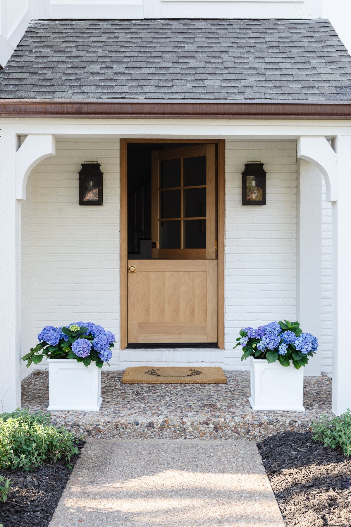 A white brick house with a wooden Dutch door and white planters on the spring porch, filled with blue hydrangeas. 