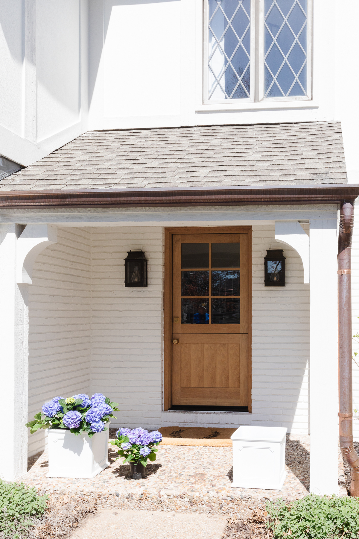 A white brick house with a wooden Dutch door and white planters on the spring porch, filled with blue hydrangeas.