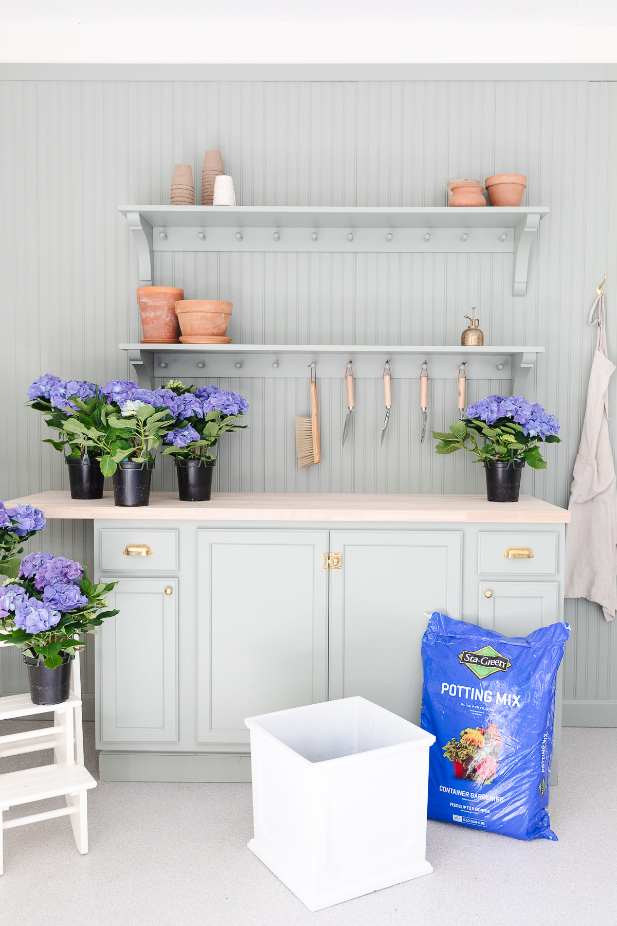 A green potting bench with a white planter and spring blue potted hydrangeas.