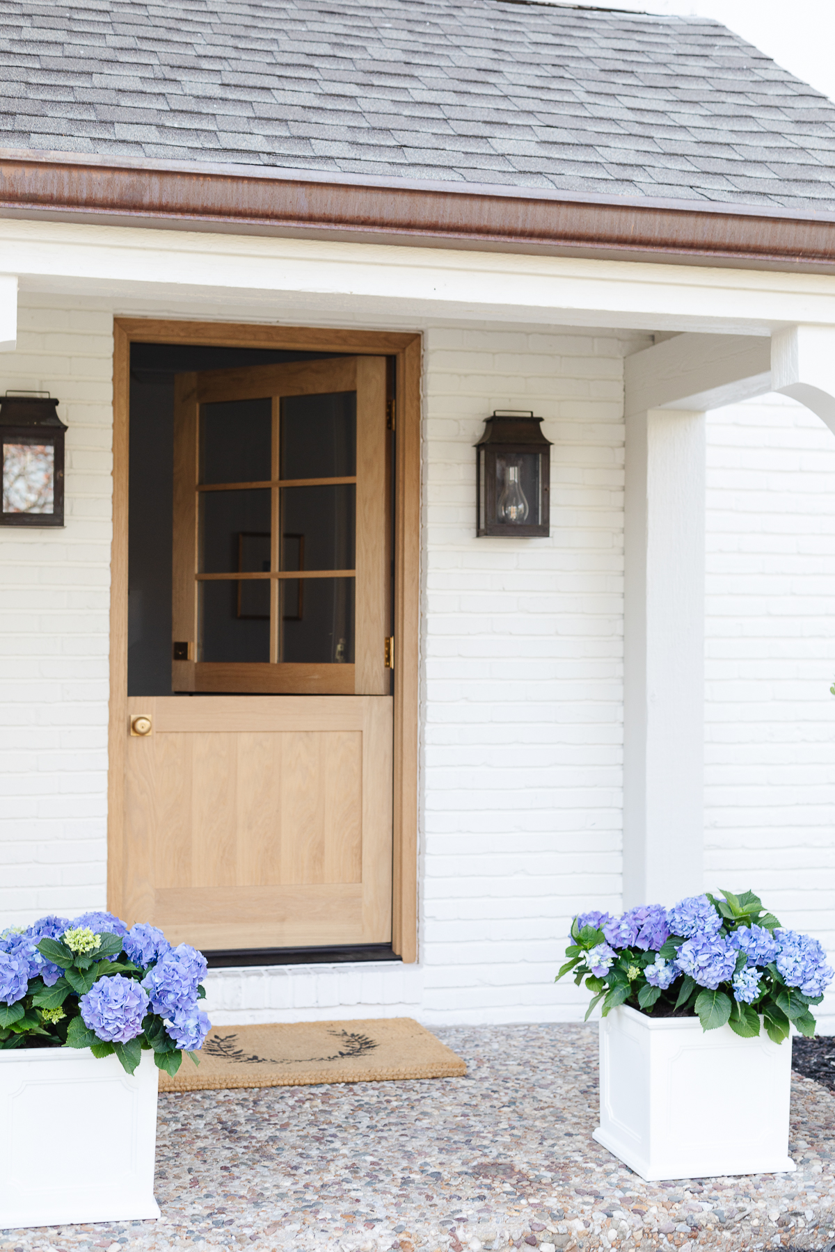 A white brick house with a wooden Dutch door and white planters on the spring porch, filled with blue hydrangeas.