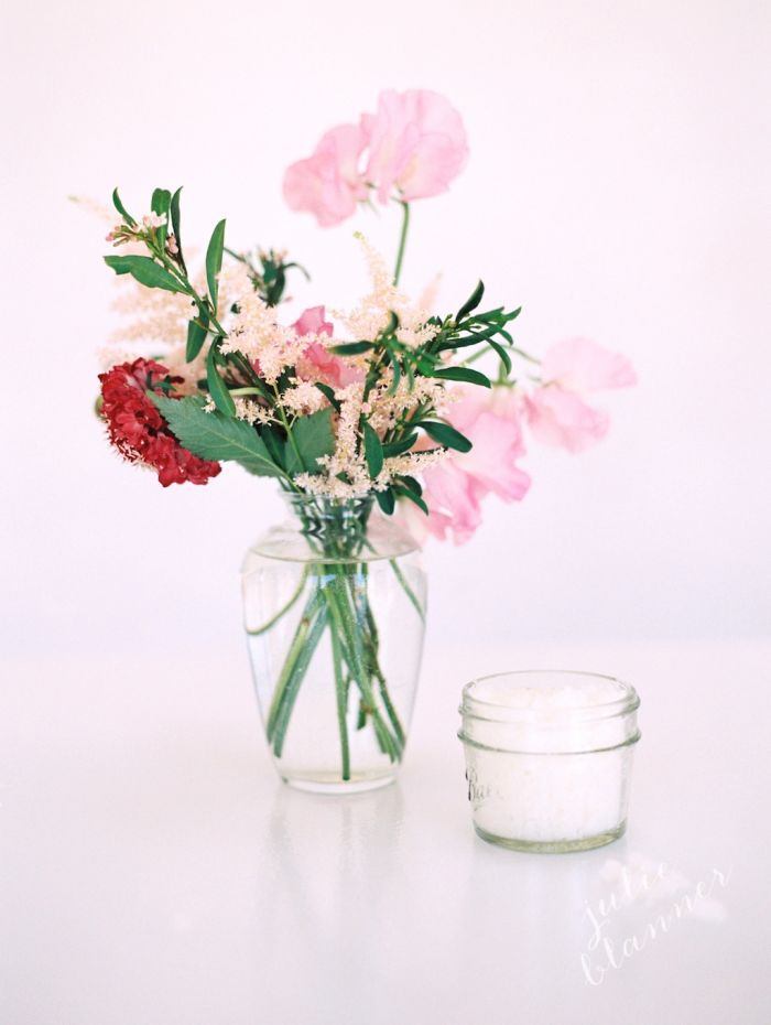 A small bouquet of flowers in a vase on a table with a jar of sugar scrub right beside it.