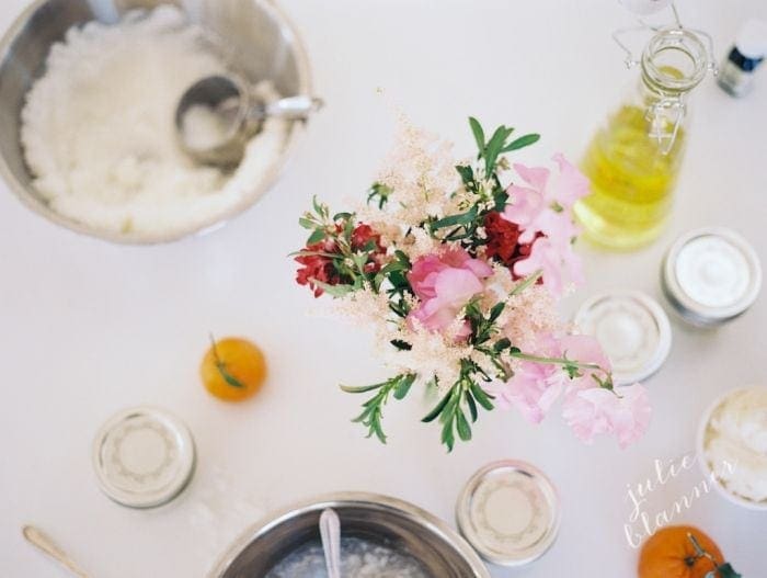 Looking down onto a scene of a  sugar scrub bar, flowers in the center and ingredients surrounding.