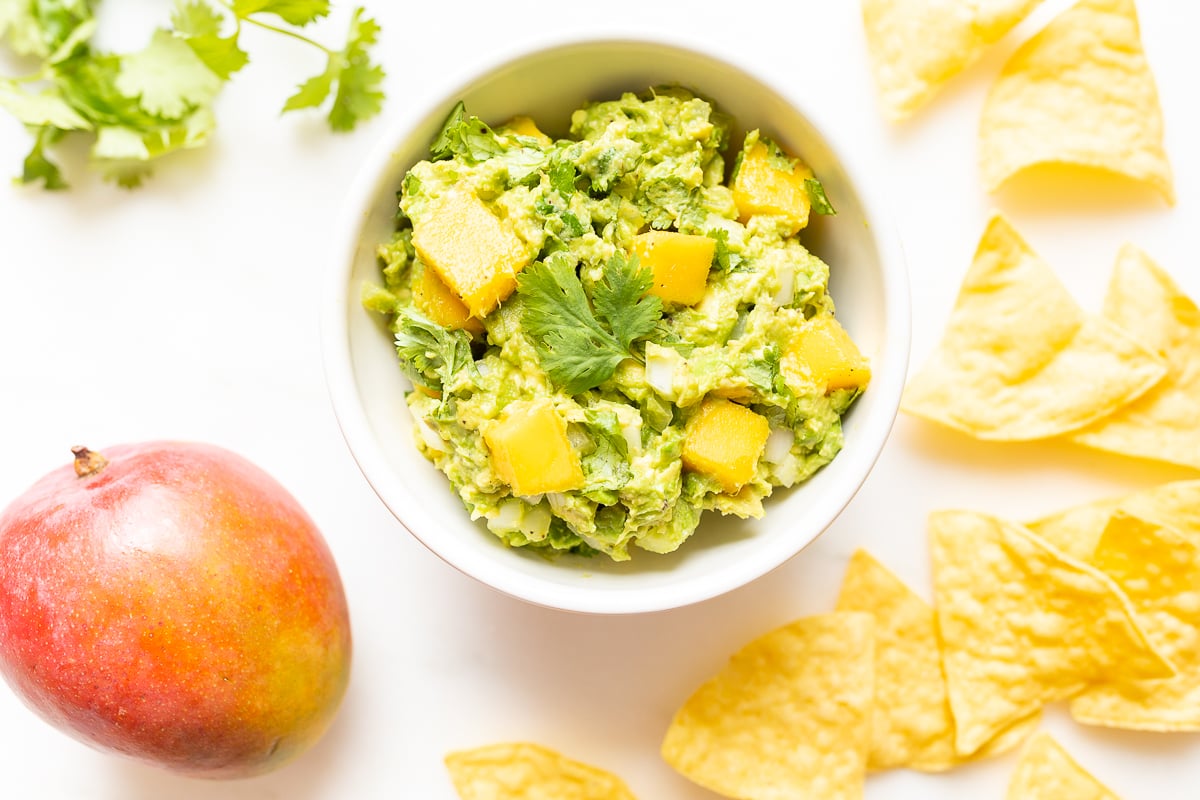 A bowl of mango guacamole surrounded by tortilla chips, along with a mango and fresh cilantro to the side.
