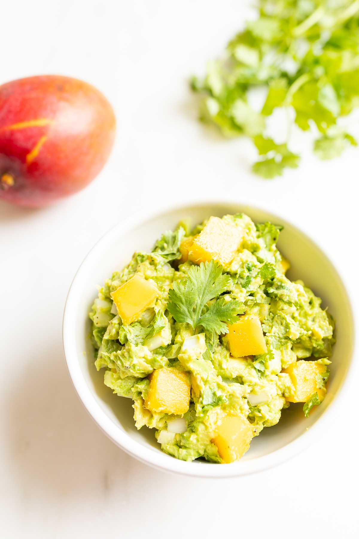 A bowl of mango guacamole surrounded by a mango and fresh cilantro to the side.