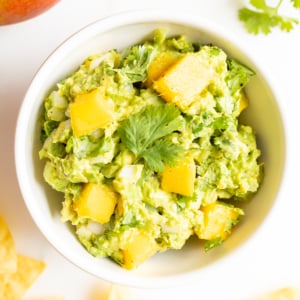 A bowl of mango guacamole surrounded by tortilla chips, along with a mango and fresh cilantro to the side.