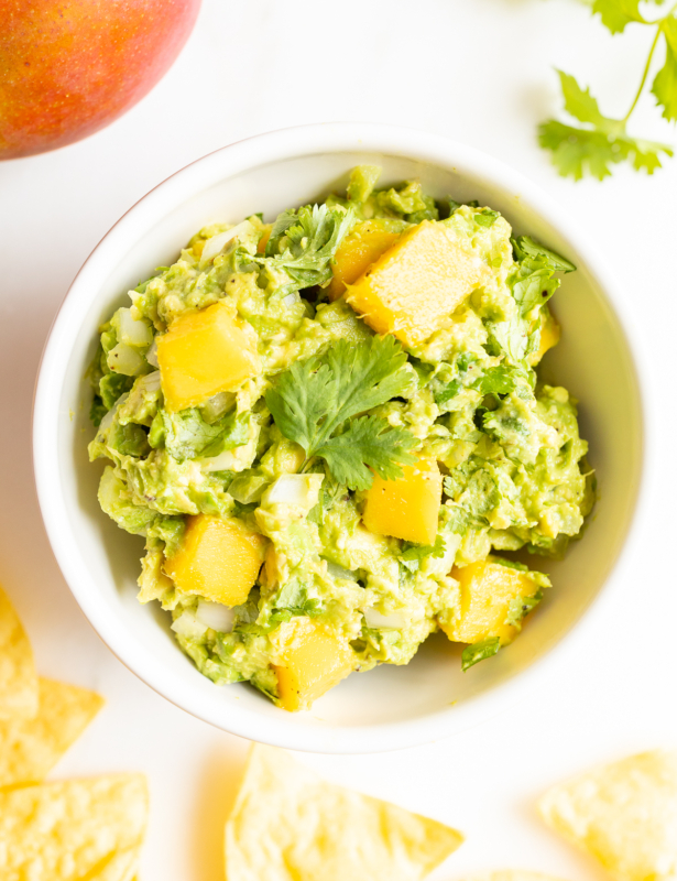 A bowl of mango guacamole surrounded by tortilla chips, along with a mango and fresh cilantro to the side.