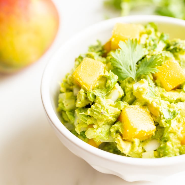 A bowl of mango guacamole surrounded by a mango and fresh cilantro to the side.