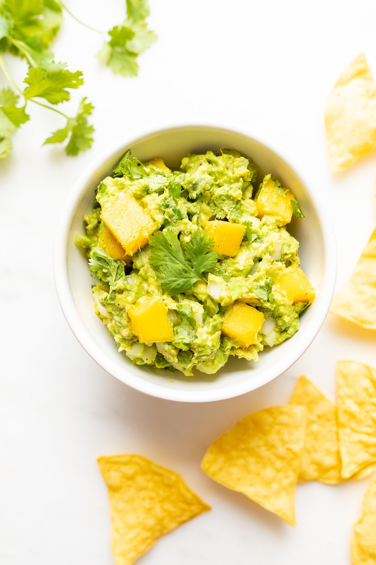 A bowl of mango guacamole surrounded by tortilla chips, along with fresh cilantro to the side.