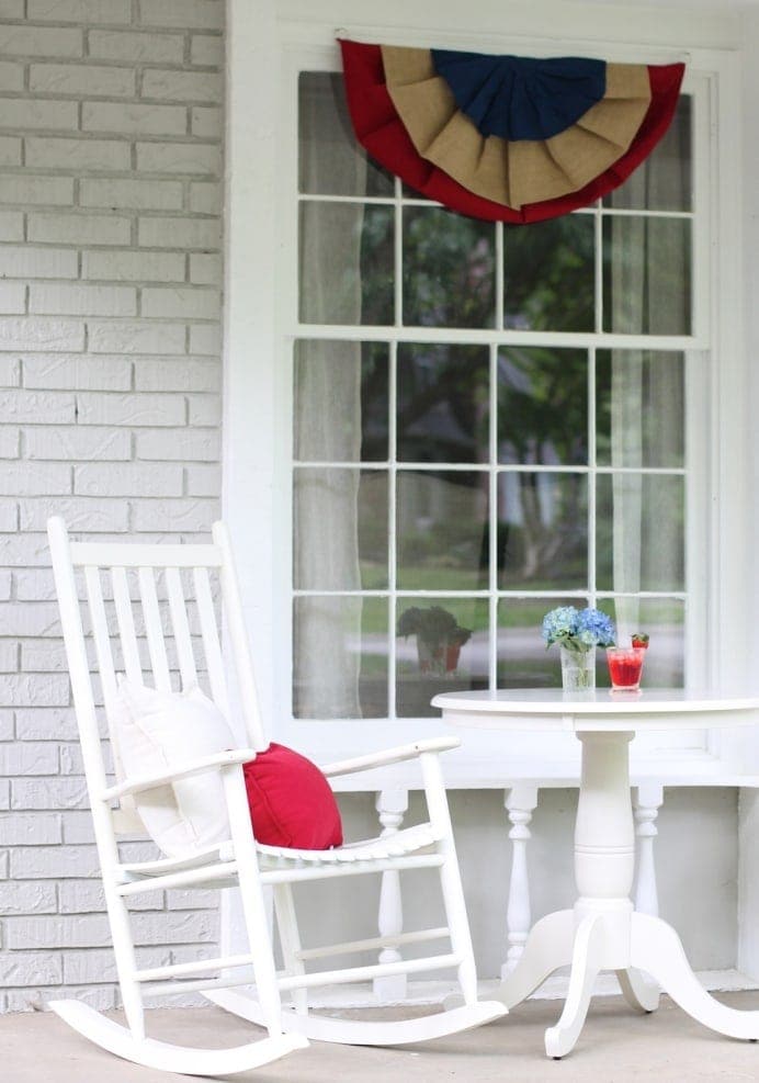 A window decoration, with a layered red, blue, and burlap decoration hanging from the top of the window. White chairs and a white table sit below.