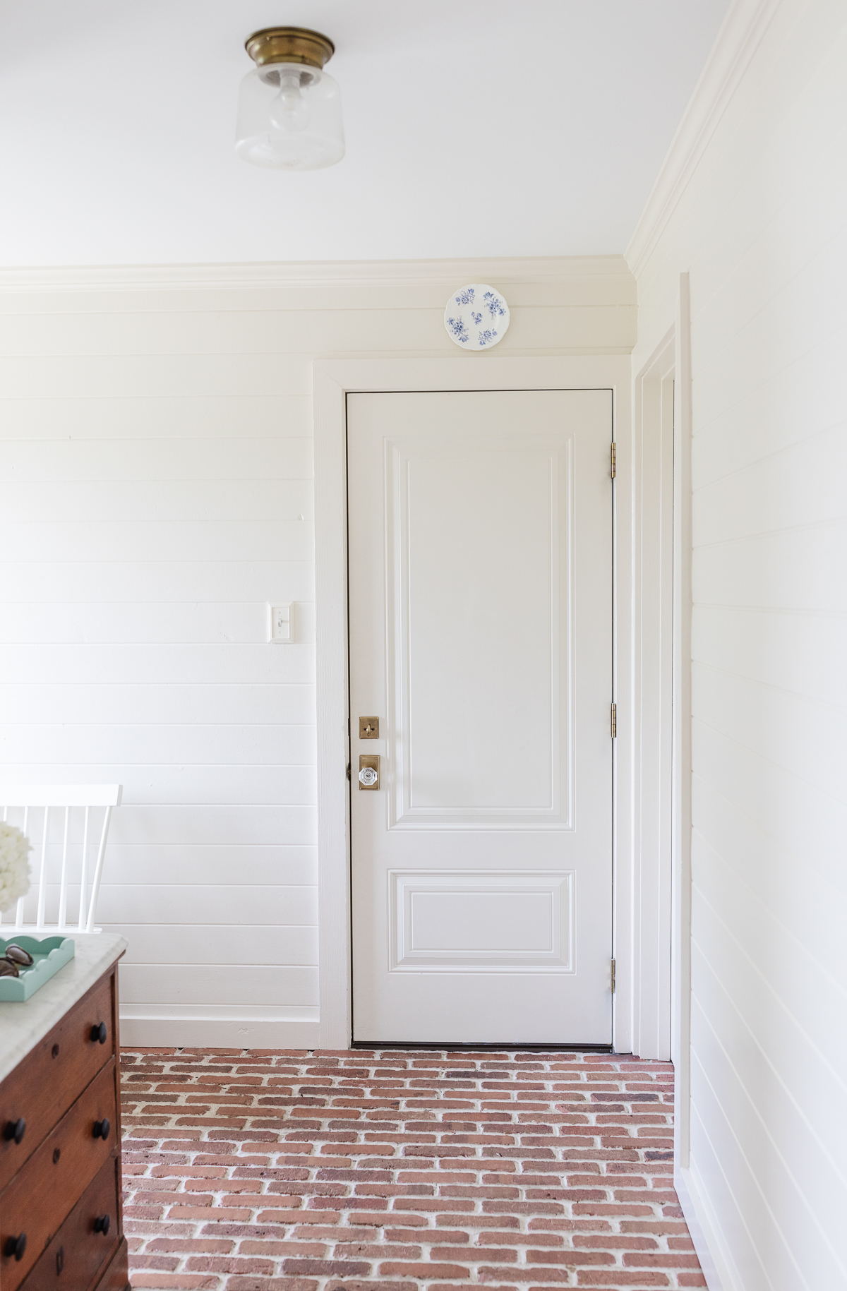 A mudroom with brick floors and paneled white walls, with a blue and white plate hanging over the door. 