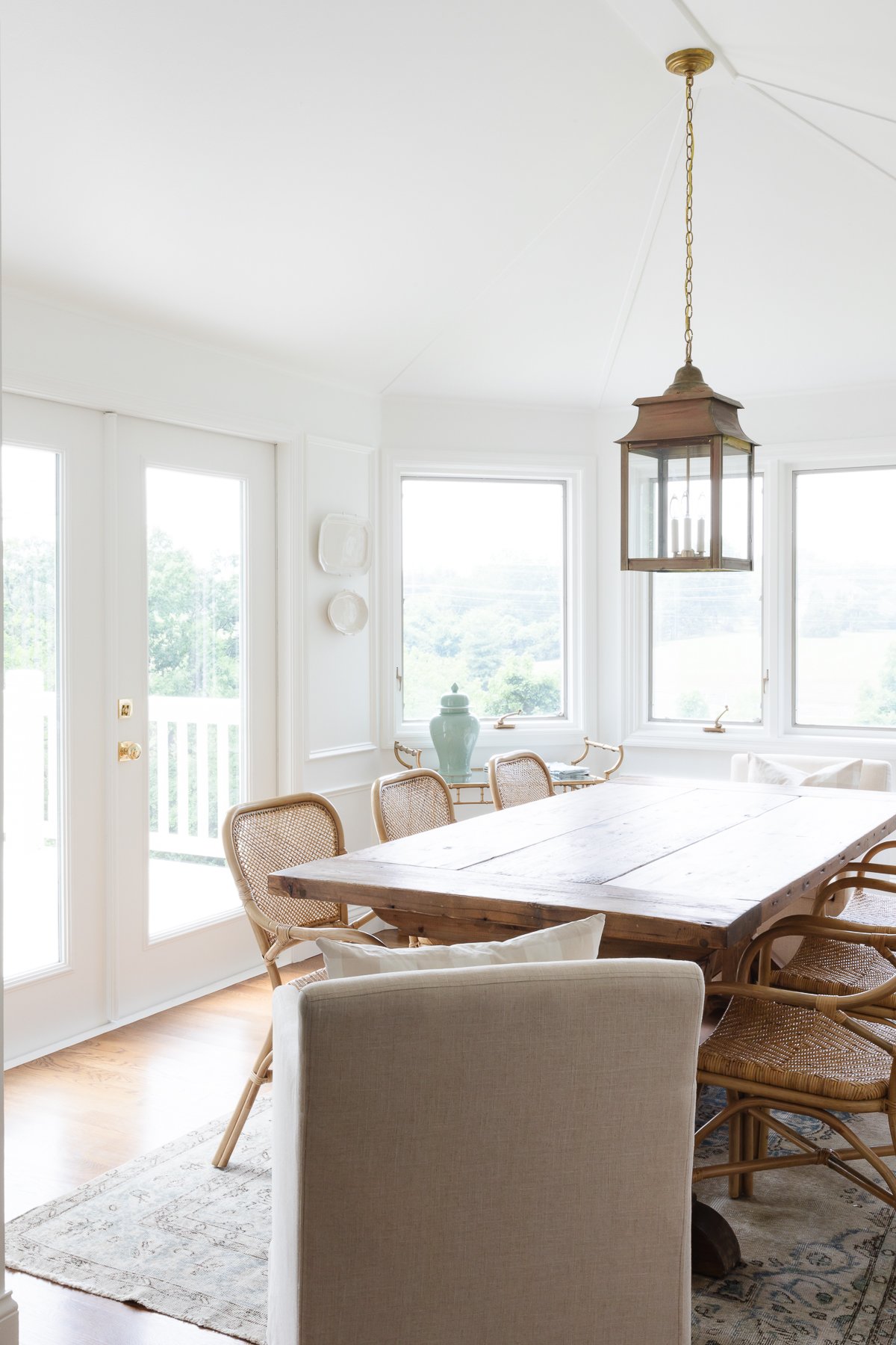 A white dining room with a wood farm table, rattan chairs, brass lantern and plates on the wall.