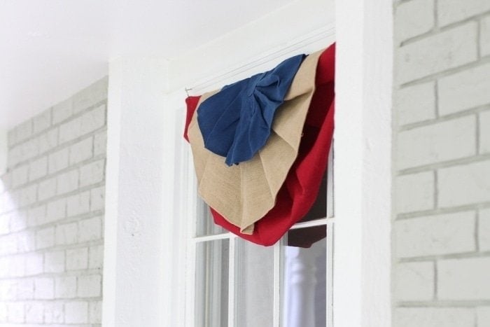 A close up of a window decoration, with a layered red, blue, and burlap decoration hanging from the top of the window. 