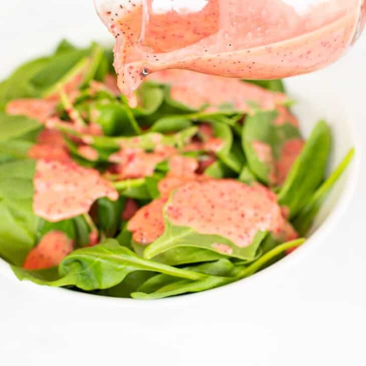 A white bowl full of fresh spinach, small glass pitcher of pink vinaigrette being poured on top.