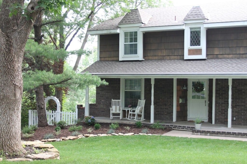A colonial home exterior in dark shingles and brick, two white rocking chairs on front porch