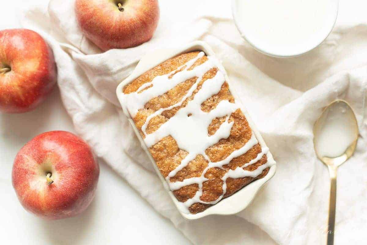 White ceramic loaf pan on a white fabric backdrop, filled with quick bread and drizzled with glaze. 