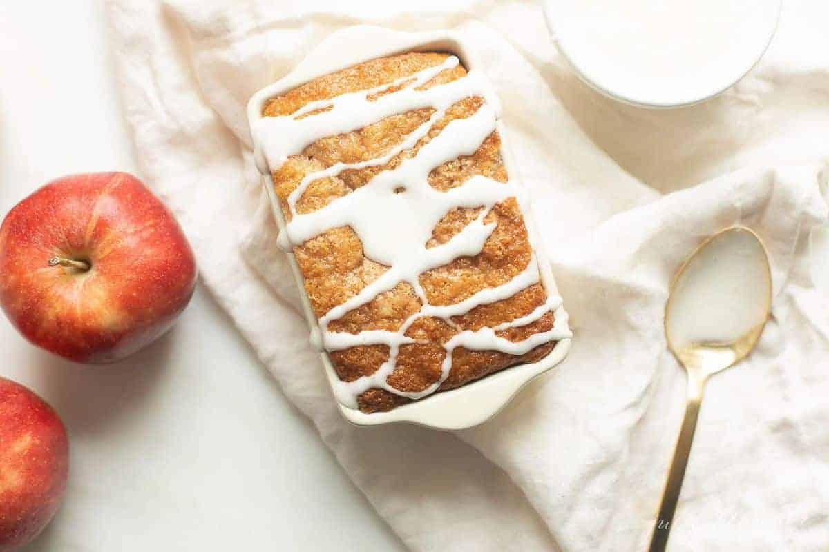 White ceramic loaf pan on a white fabric backdrop, filled with quick bread and drizzled with glaze. 