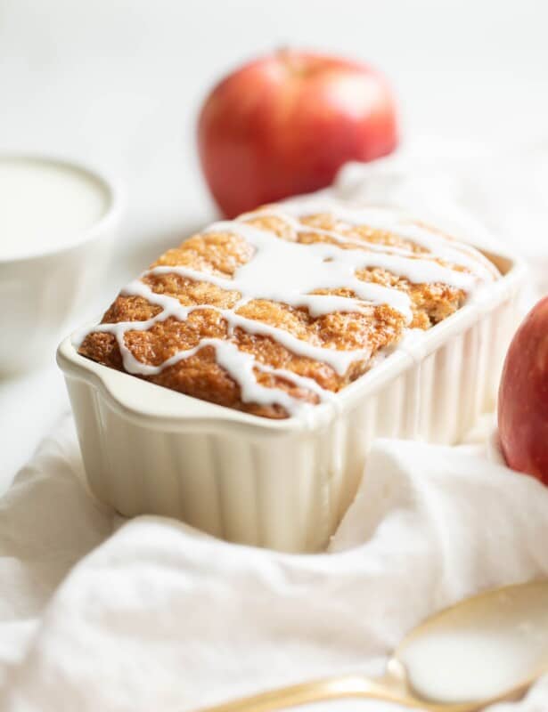 White ceramic loaf pan on a white fabric backdrop, filled with apple bread and drizzled with glaze.