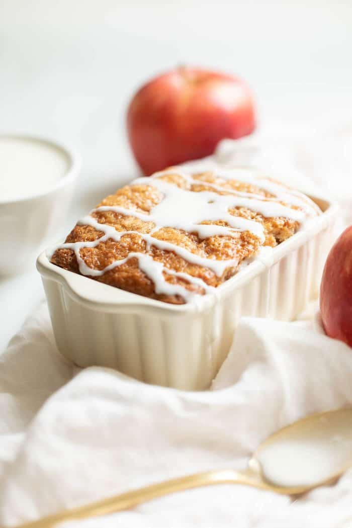 White ceramic loaf pan on a white fabric backdrop, filled with apple bread and drizzled with glaze.