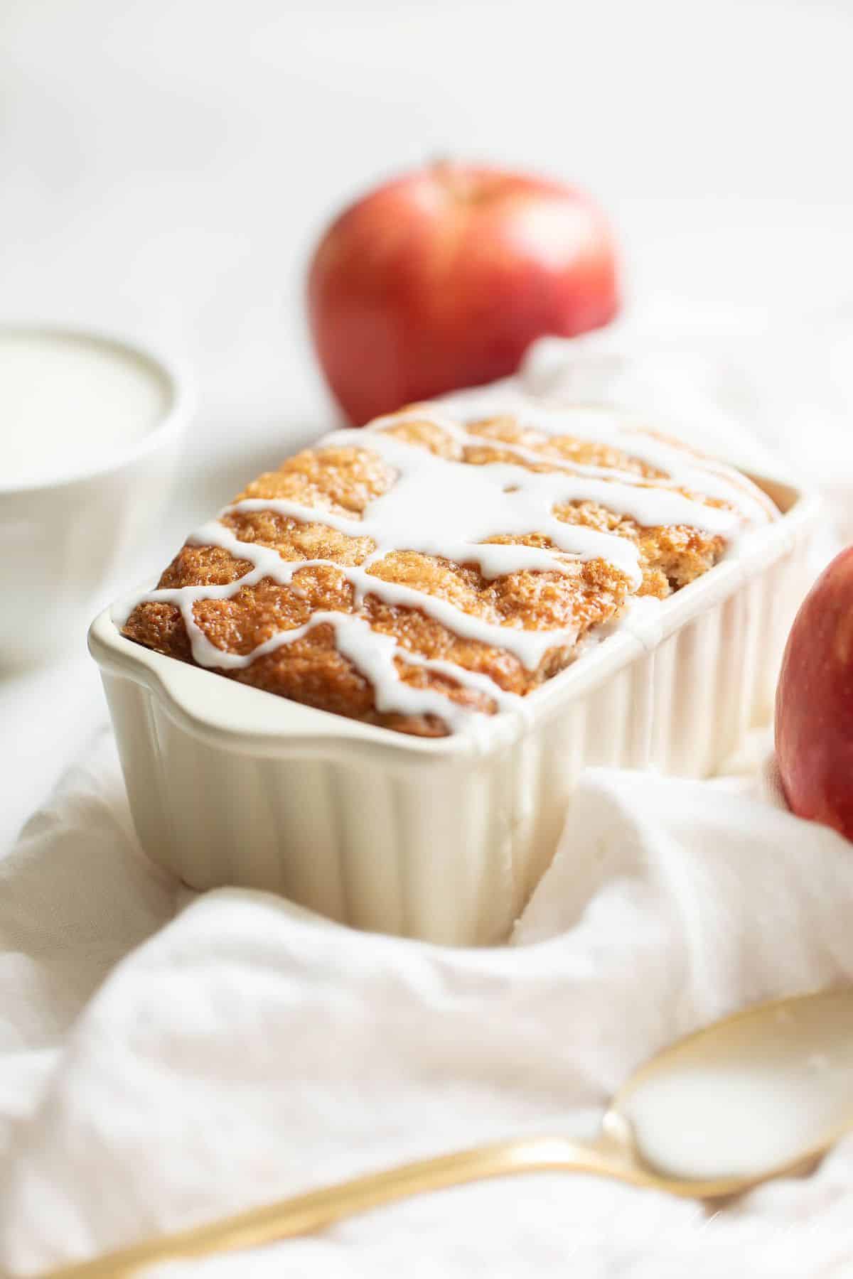 White ceramic loaf pan on a white fabric backdrop, filled with quick bread and drizzled with glaze. 