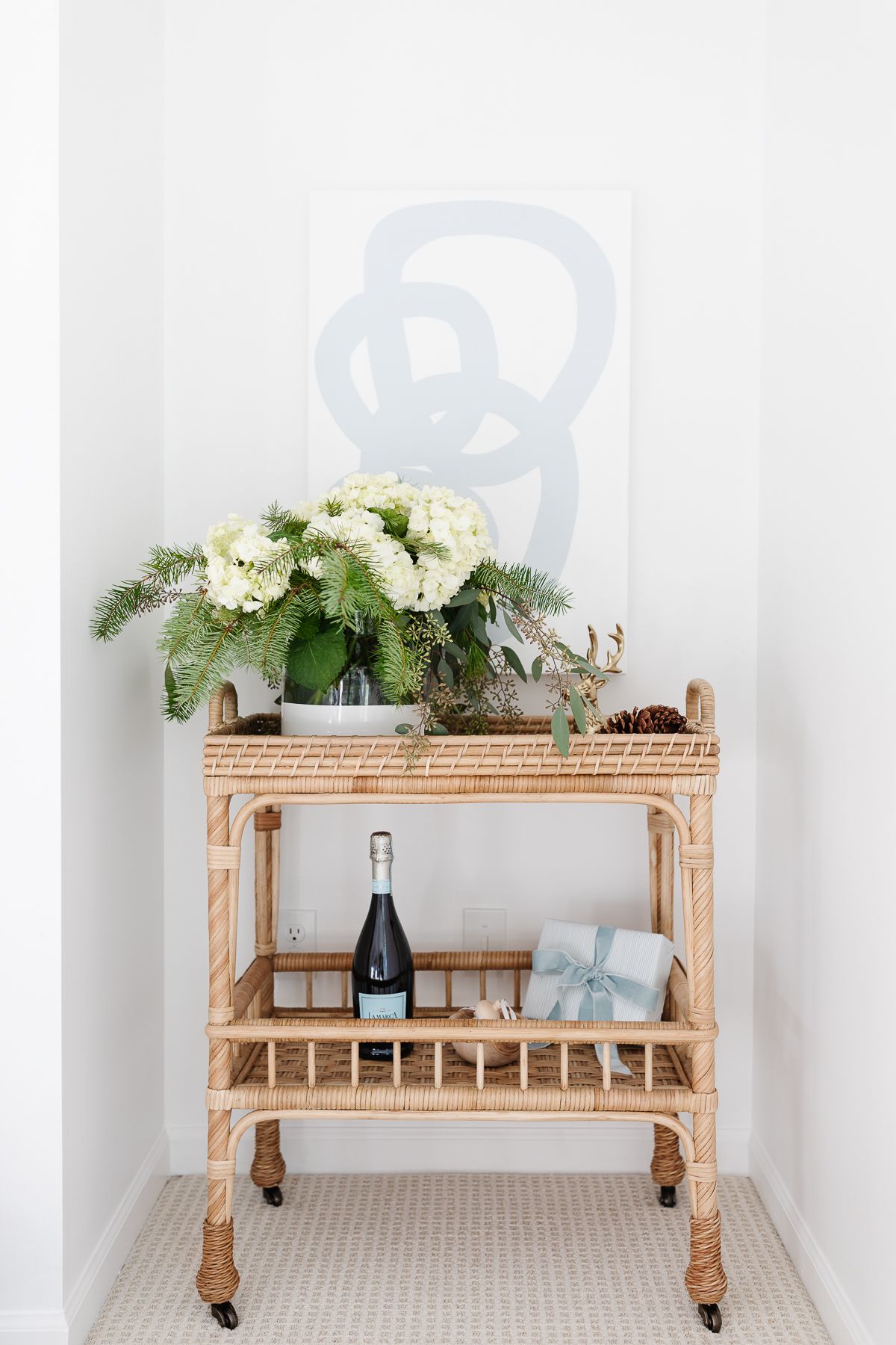 A rattan bar cart styled for Christmas with a vase of fresh flowers and modern art behind it.