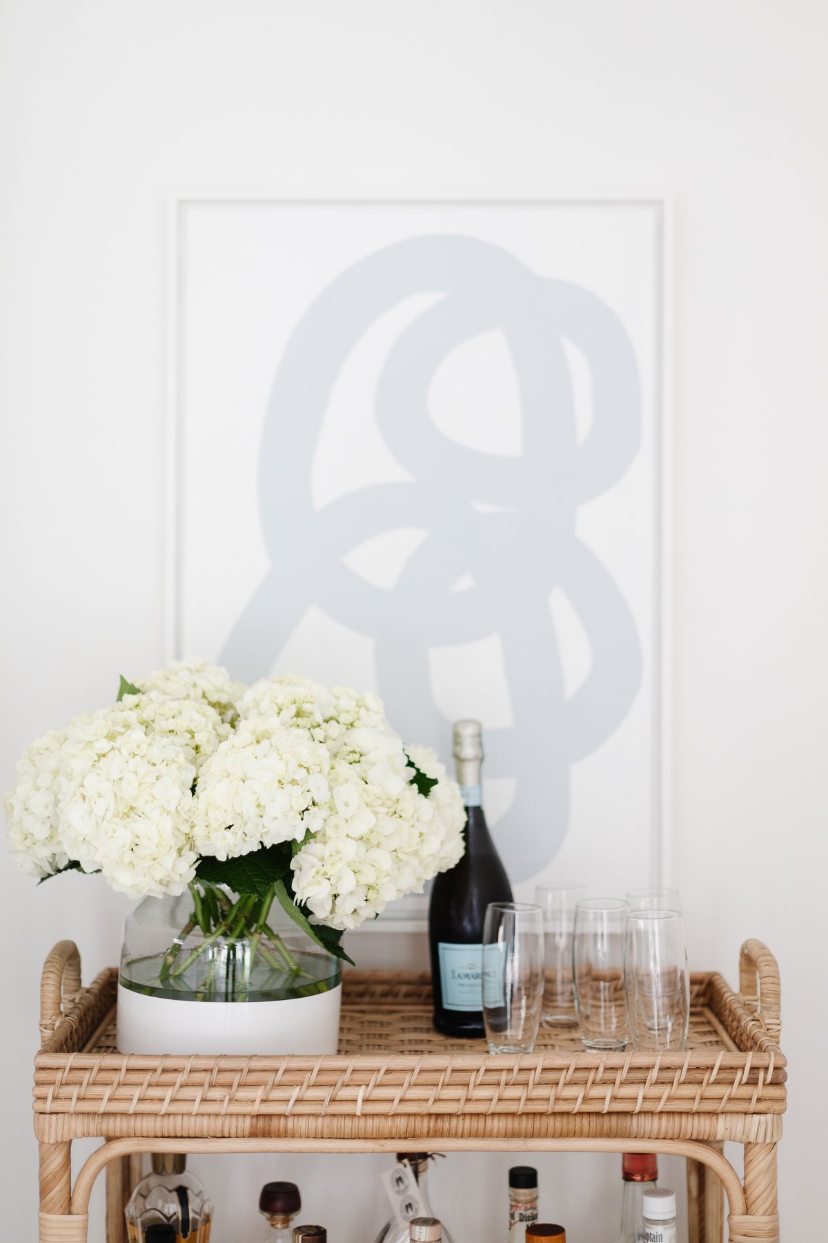 A rattan bar cart with a vase of fresh flowers and modern art behind it.