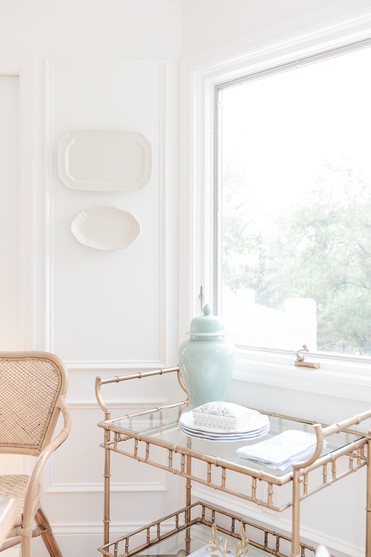 A gold bar cart styled with blue and white accessories in a white dining room. Rattan chair nearby.