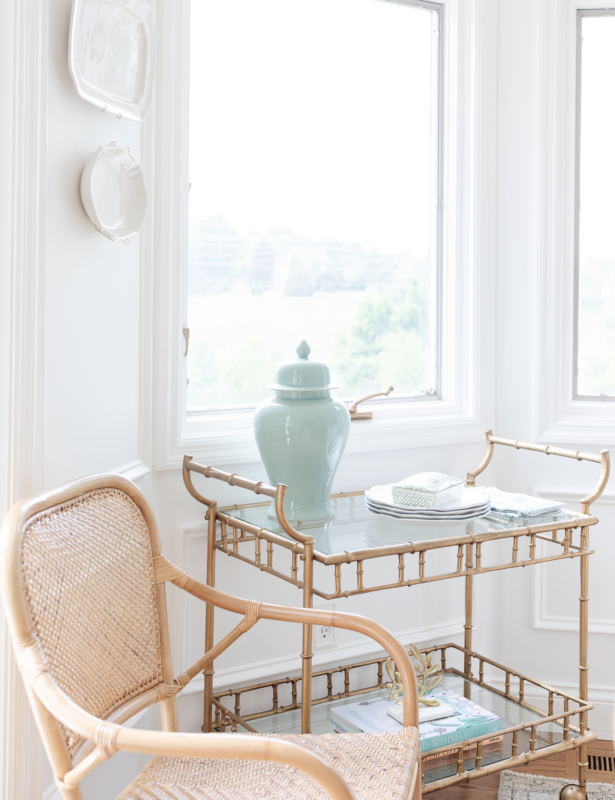 A gold bar cart styled with blue and white accessories in a white dining room. Rattan chair nearby.