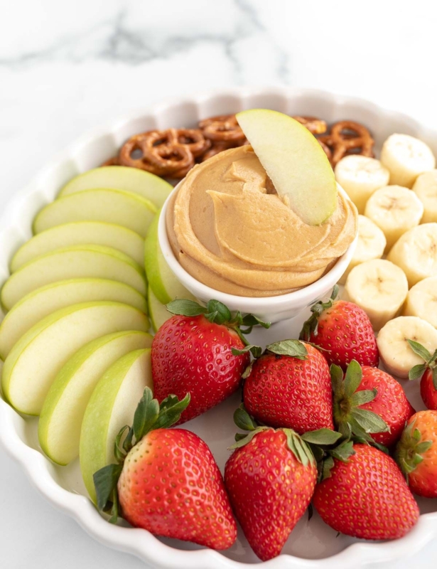 A white platter, with peanut butter dip in the center, surrounded by fruit and pretzels.