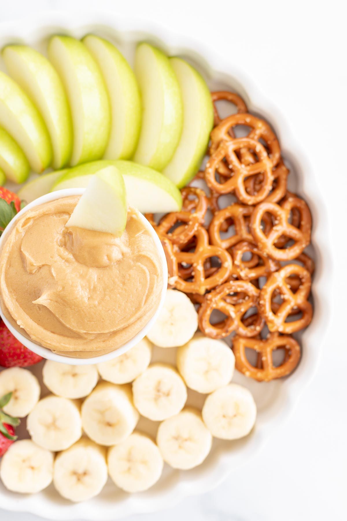 A white platter, with peanut butter fruit dip in the center, surrounded by fruit and pretzels.