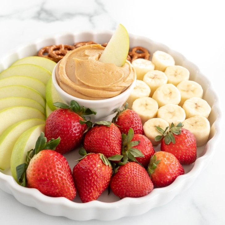A white platter, with peanut butter dip in the center, surrounded by fruit and pretzels.