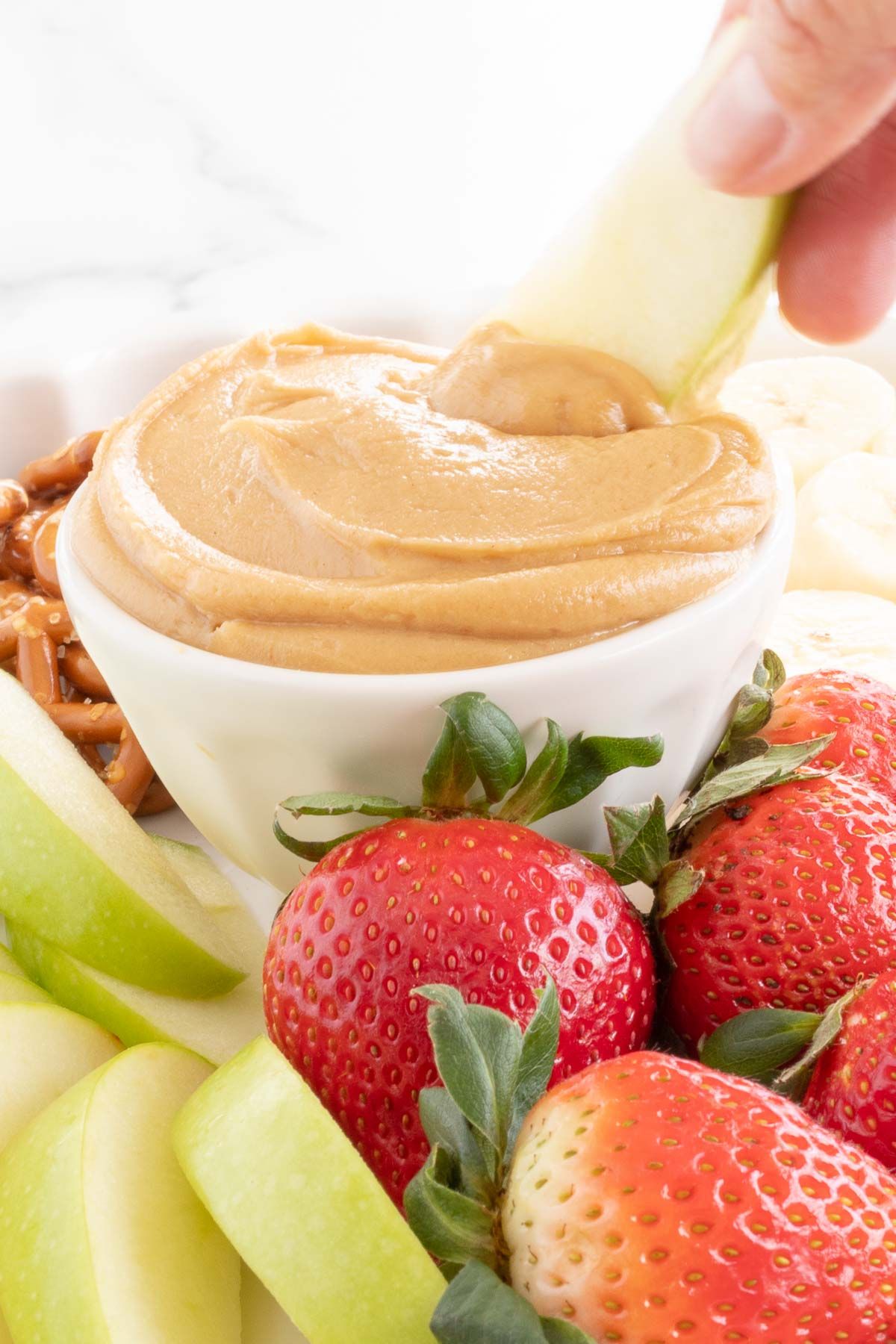 A white platter, with peanut butter fruit dip in the center, surrounded by fruit and pretzels.