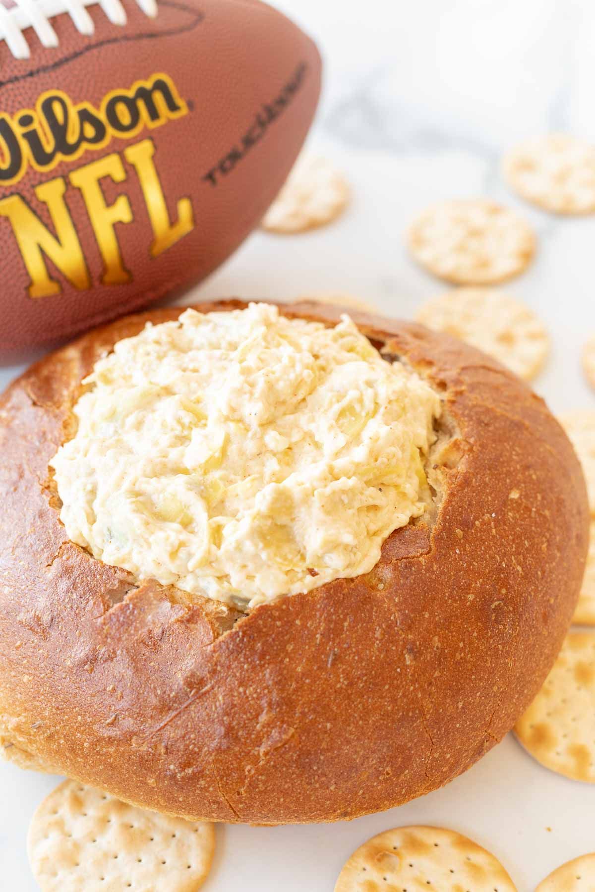 A bread bowl full of a spinach dip recipe with a football in the background.