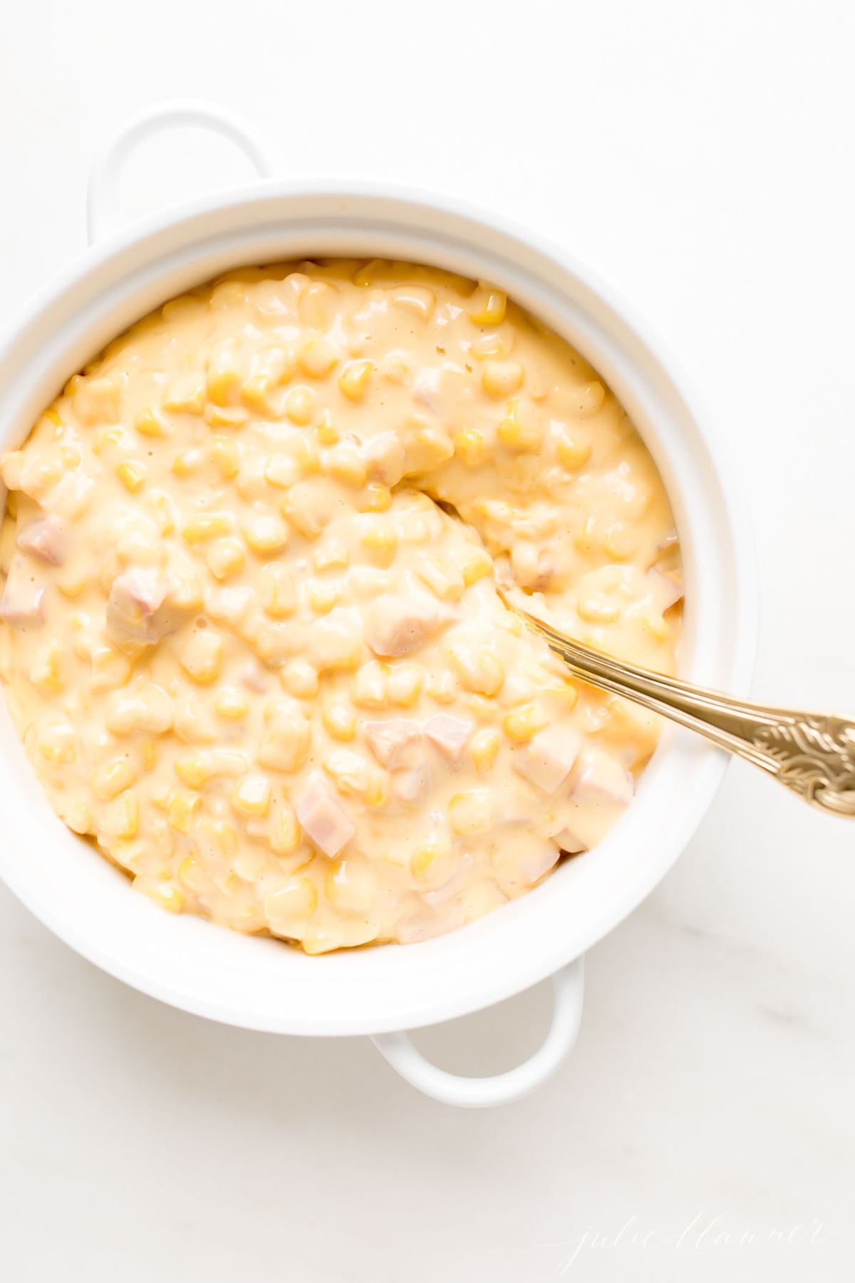 A white casserole dish filled with Cheesy Corn Casserole, a gold serving spoon in the center.