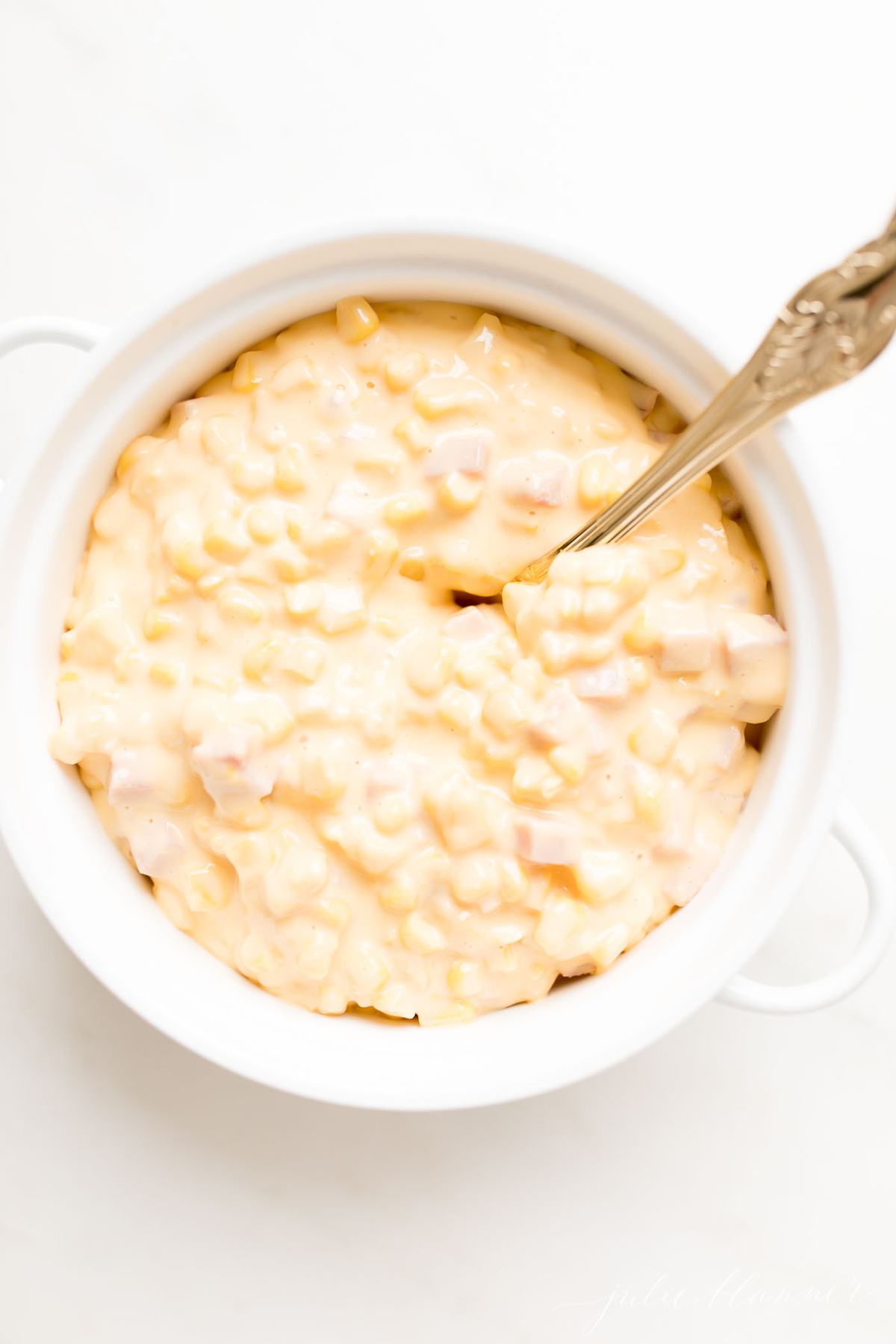 A white casserole dish filled with Cheesy Corn Casserole, a gold spoon in the center.