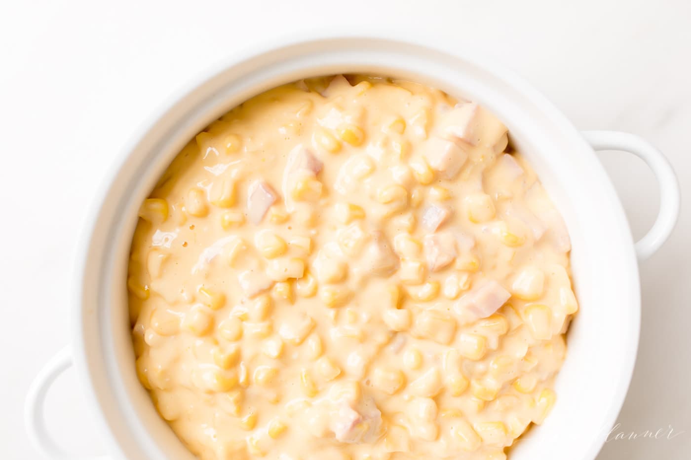 A white casserole dish with handles, filled with cheesy corn dip with ham.